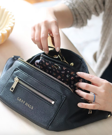 A black belt bag is partially open, revealing a floral lining. A hand is reaching into the bag. The bag features two zippered compartments on the front, and the words